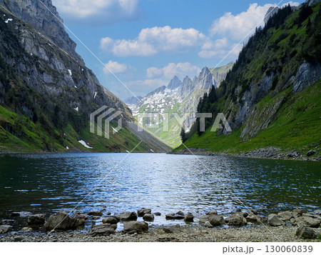 Scenic mountain lake surrounded by green hills and rocky cliffs under a blue sky with clouds 130060839