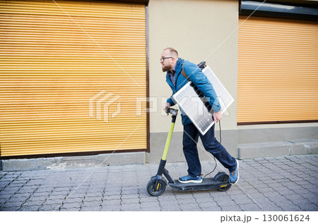 Man rides electric scooter while carrying solar panel. Integration of solar power as sustainable energy source for charging electric scooters, promoting eco-friendly urban transportation. Man rides electric scooter while carrying solar panel. Integration of solar power as sustainable energy source for charging electric scooters, promoting eco-friendly urban transportation. 130061624
