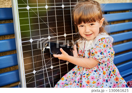 Young girl holds smartphone connected to solar panel for charging. Integration of sustainable renewable energy into daily life, importance of teaching children about eco-friendly technology. 130061625