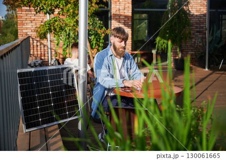 Man works on laptop and smartphone at outdoor table. Solar panel nearby, emphasizing sustainable, modern eco-friendly workspace that combines technology and sustainability. Man works on laptop and smartphone at outdoor table. Solar panel nearby, emphasizing sustainable, modern eco-friendly workspace that combines technology and sustainability. 130061665