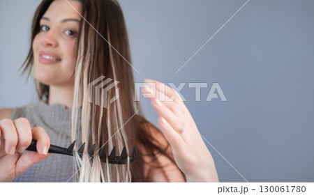 Young woman with hair extensions gently combing hair using comfortable safe comb in beauty salon 130061780