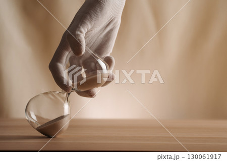 A glove-covered hand holding a tipped hourglass on a wooden surface, symbolizing the flow of time, creativity, and patience in a contemporary minimalist style. A glove-covered hand holding a tipped hourglass on a wooden surface, symbolizing the flow of time, creativity, and patience in a contemporary minimalist style. 130061917