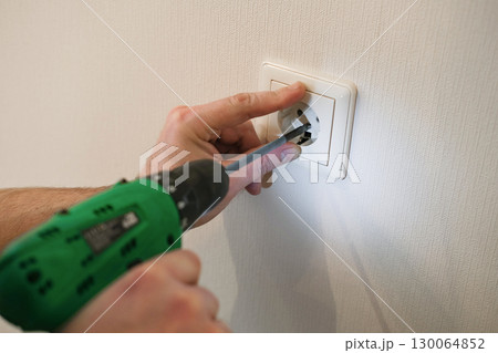 Person uses a power screwdriver to install electrical outlet cover plate on white wall. Home repair, DIY projects, electrical work and professional craftsmanship 130064852