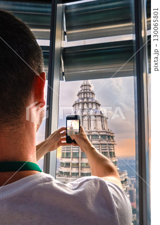 Nov 20, 2019 - Kuala Lumpur, Malaysia: Unrecognizable male adult tourist taking photo of Petronas tower with smartphone through window at colorful sunset.Observation deck visit, tourist attraction Nov 20, 2019 - Kuala Lumpur, Malaysia: Unrecognizable male adult tourist taking photo of Petronas tower with smartphone through window at colorful sunset.Observation deck visit, tourist attraction 130065801