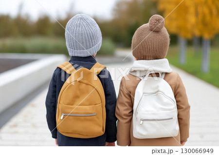 Two children wearing cozy knit hats and backpacks stand on a path surrounded by autumn trees 130066949