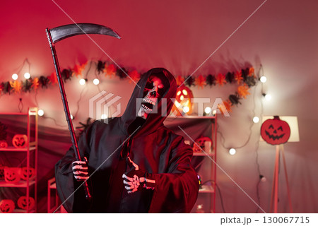 Grim reaper holding scythe in halloween decorated room Grim reaper holding scythe in halloween decorated room 130067715