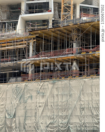 Modern building under construction with visible scaffolding, yellow framework, concrete columns, and protective green mesh netting, active urban development Modern building under construction with visible scaffolding, yellow framework, concrete columns, and protective green mesh netting, active urban development 130067802