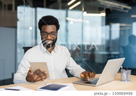 Professional individual using tablet and laptop at office desk with focused expression in corporate environment. 130069498