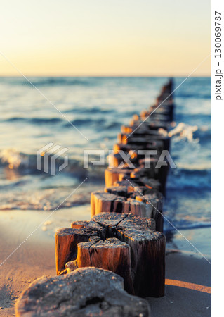 Wooden breakwater stretches into blue sea under soft evening light. Gentle waves touch sandy shore while sun reflects warmly on weathered timber posts and shimmering water Wooden breakwater stretches into blue sea under soft evening light. Gentle waves touch sandy shore while sun reflects warmly on weathered timber posts and shimmering water 130069787
