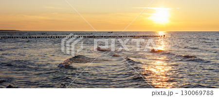 Scenic warm sea sunset view golden sunset over Baltic Sea reflects across rippling waves, creating a glowing path of light. Wooden breakwaters stretch into horizon calm rhythm to tranquil seascape 130069788