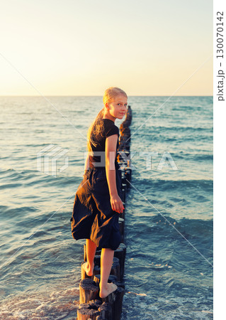 Cute adorable blond teenager happy kid girl walking by wooden breakwater posts at Baltic sea scenic warm summer sunset light. Balancing reflects challenges, choices and uncertainties of growing up 130070142