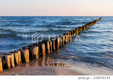 Wooden breakwater stretches into blue sea under soft evening light. Gentle waves touch sandy shore while sun reflects warmly on weathered timber posts and shimmering water 130070143