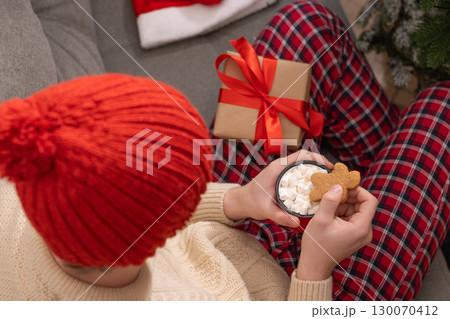 Young man in red plaid pajamas holding mug of hot cocoa topped with marshmallows and a gingerbread cookie. Festive atmosphere with warm lights and red beanie hat enhancing coziness. Xmas holiday. Home Young man in red plaid pajamas holding mug of hot cocoa topped with marshmallows and a gingerbread cookie. Festive atmosphere with warm lights and red beanie hat enhancing coziness. Xmas holiday. Home 130070412
