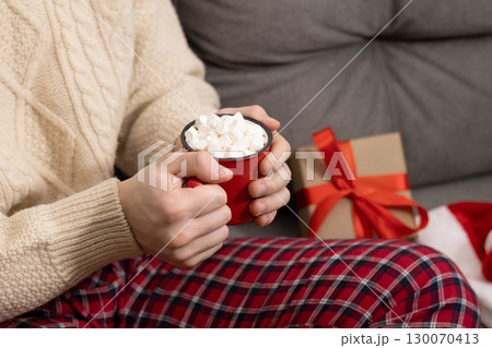Christmas coffee. Young man in red plaid pajamas holding mug of hot cocoa topped with marshmallows. Festive holiday atmosphere. Xmas holiday. Home interior. Christmas coffee. Young man in red plaid pajamas holding mug of hot cocoa topped with marshmallows. Festive holiday atmosphere. Xmas holiday. Home interior. 130070413