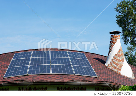 Solar panels installed on a curved red roof with chimney under blue sky, renewable energy concept 130070750