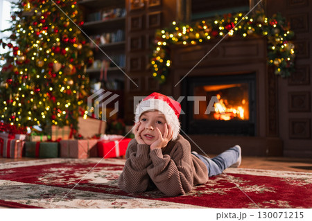 Merry Christmas. Little boy smiling near Christmas tree in classical dark interior. Young happy kid wearing Santa Hat in living room with fireplace Christmas tree gift boxes. Christmas eve at home 130071215