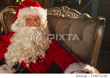 Merry Christmas. Happy Santa Claus sitting in chair in festive living room with fireplace decorations Christmas tree. Santa in cozy home on Christmas eve. Winter New Year Holiday Celebrations 130071248