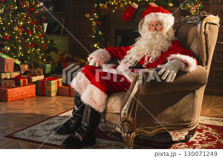 Merry Christmas. Happy Santa Claus sitting in chair in festive living room with fireplace decorations Christmas tree. Santa in cozy home on Christmas eve. Winter New Year Holiday Celebrations 130071249
