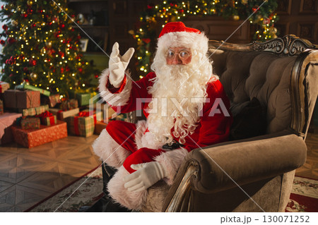 Merry Christmas. Happy Santa Claus sitting in chair in festive living room with fireplace decorations Christmas tree. Santa in cozy home on Christmas eve. Winter New Year Holiday Celebrations 130071252