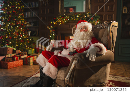 Merry Christmas. Happy Santa Claus sitting in chair in festive living room with fireplace decorations Christmas tree. Santa in cozy home on Christmas eve. Winter New Year Holiday Celebrations 130071253