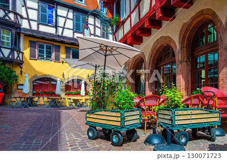 Street restaurant outdoor cafe tables, chairs and umbrellas, old colorful building fachwerk style facade and shutters windows in old town Colmar city historic centre, Alsace Grand Est region, France 130071723