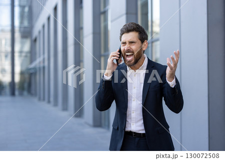A businessman in a formal suit angrily reacts during a phone call near an office. His emotions indicate frustration, highlighting conflict in a professional setting A businessman in a formal suit angrily reacts during a phone call near an office. His emotions indicate frustration, highlighting conflict in a professional setting 130072508