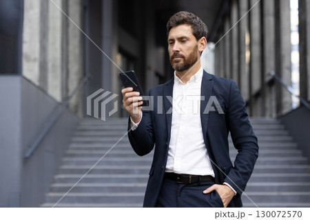 A professional businessman checks his smartphone on a modern urban staircase, dressed in a formal suit, showcasing confidence and connectivity in a professional environment. 130072570