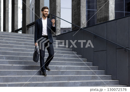 Businessman wearing a formal suit using his smartphone, walking, and carrying a bag outdoors, showcasing professionalism, connectivity, and a vibrant urban environment 130072576