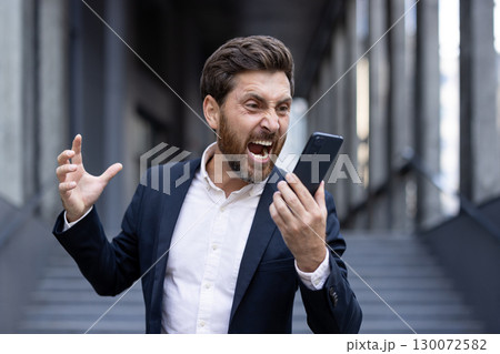 A professionally dressed man visibly shouting in frustration at his smartphone in an urban environment. His expression conveys deep anger or annoyance A professionally dressed man visibly shouting in frustration at his smartphone in an urban environment. His expression conveys deep anger or annoyance 130072582