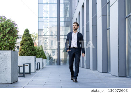 A confident man in a suit walks through an urban office complex, surrounded by greenery. A confident man in a suit walks through an urban office complex, surrounded by greenery. 130072608