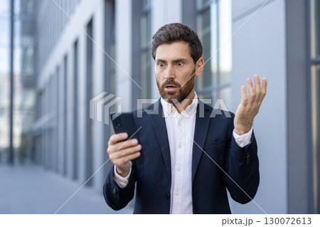 A professional man in a suit expressing shock while looking at his phone. The background showcases modern architecture, emphasizing the urban setting and suggesting a concept of unexpected news A professional man in a suit expressing shock while looking at his phone. The background showcases modern architecture, emphasizing the urban setting and suggesting a concept of unexpected news 130072613