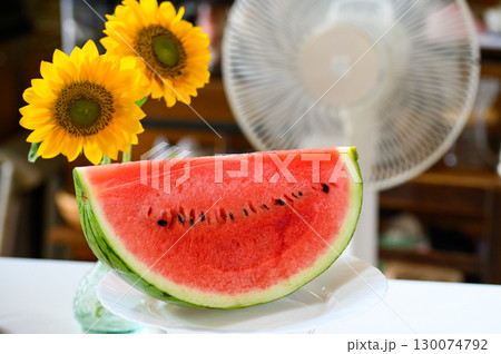 室内のテーブルに置かれた切ったスイカと風に揺れるひまわりと回る扇風機 室内のテーブルに置かれた切ったスイカと風に揺れるひまわりと回る扇風機 130074792