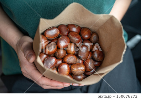 Cropped shot view of someone holding roasted chestnut in paper bag. Roasted chestnut is a popular autumn and winter street food in Asia. Cropped shot view of someone holding roasted chestnut in paper bag. Roasted chestnut is a popular autumn and winter street food in Asia. 130076272