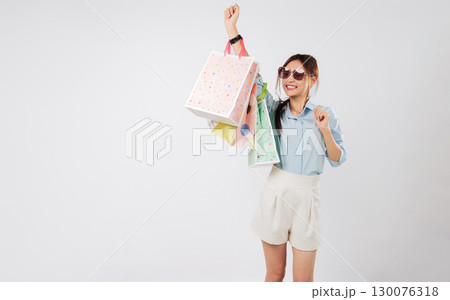 Happy young Asian woman shopping, raising hand, carrying paper bags after Black Friday, she excited and having fun dancing with a winner gesture holding her shopping bags, isolated white background 130076318