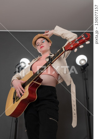 Portrait of confident female guitarist holding guitar, looking at camera. With one hand she holds brim of her straw hat, her stage outfit complemented by leather jacket and jeans. Low angle view 130077157