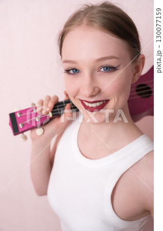 Young female musician holding ukulele by neck and slinging musical instrument over shoulder, she's feeling inspired. Beautiful woman looking camera with smile, positive attitude and happy expression 130077159