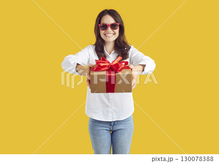 Cheerful woman with joyful expression hands you festive gift box tied with red ribbon. 130078388