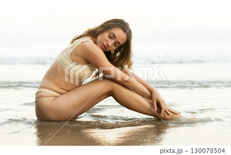 Young girl in bikini panties and bra sitting on sandy seashore. Young girl in bikini panties and bra sitting on sandy seashore. 130078504