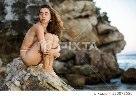 Topless girl in panties posing near sea, model sitting on large boulder 130078586