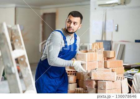 Builder in blue workwear planning erection of partition wall in building under reconstruction, preparing red bricks for work 130078646