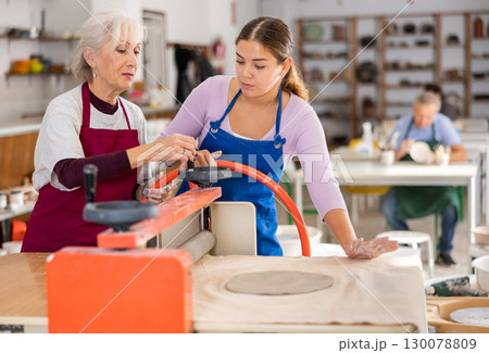 Experienced elderly woman potter teaches young student how to work on craft machine clay press roller Experienced elderly woman potter teaches young student how to work on craft machine clay press roller 130078809