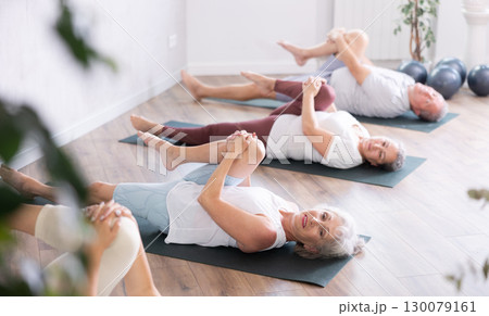 Class of elderly sporty women stretching gluteal muscle while lying on back during yoga Pilates workout on mats in gym studio 130079161