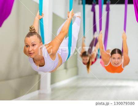 Full length side view portrait of woman hanging from silk hammock. Girl in elegant position practicing aerial yoga Full length side view portrait of woman hanging from silk hammock. Girl in elegant position practicing aerial yoga 130079380