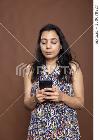 Indian woman using mobile device with focused expression, checking her emails and chats. Serious female individual standing and texting on smartphone against brown background studio. 130079827