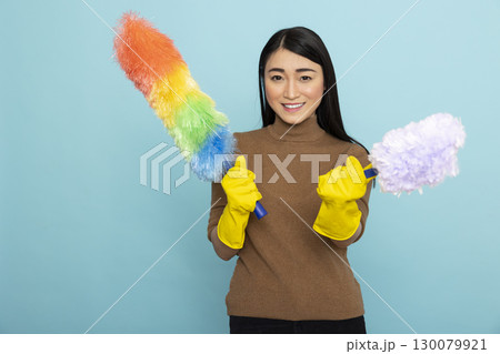 Beautiful female cleaner grasping rainbow and white duster and looking at camera. Portrait of asian housekeeper holding dusting supplies, promoting cleaning services against isolated background. 130079921