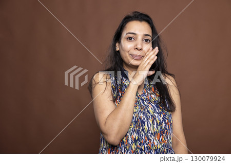 Smiling Indian woman cupping her mouth, leaning in with playful expression as she whispers a secret. Closeup portrait of female individual, conveying curiosity, confidentiality and personal moment. Smiling Indian woman cupping her mouth, leaning in with playful expression as she whispers a secret. Closeup portrait of female individual, conveying curiosity, confidentiality and personal moment. 130079924