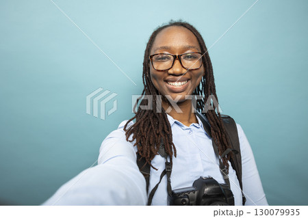 POV of trendy black woman grinning at camera while filming herself in a studio. African american female photographer stands with DSLR camera resting on her neck as she documents her creative process. 130079935