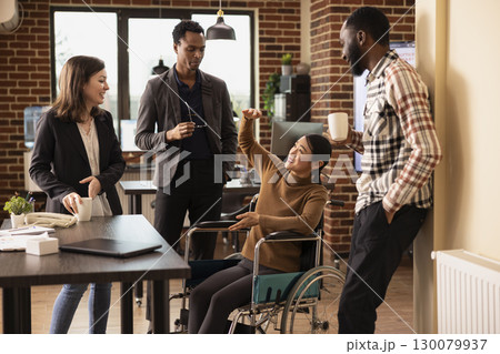 Professional employees having a lighthearted conversation, promoting teamwork in a business setting. Cheerful asian woman in a wheelchair chatting with her inclusive team, taking a short break. 130079937
