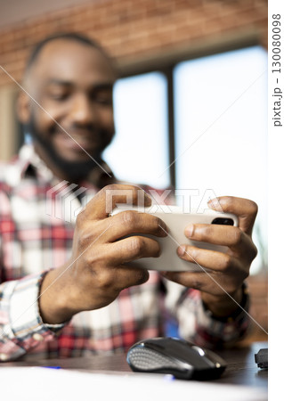 African american male entrepreneur resting from work, playing mobile games on digital device. Young adult freelancer enjoying fun and entertainment at desk in cozy apartment office. African american male entrepreneur resting from work, playing mobile games on digital device. Young adult freelancer enjoying fun and entertainment at desk in cozy apartment office. 130080098