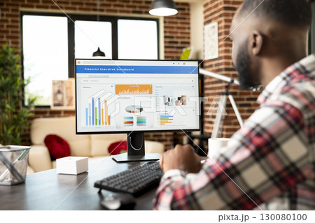 Black businessman looking at desktop monitor, studying company financial documents at home office. Remote worker analyzes business performance and prepares strategic growth plans for startup success. Black businessman looking at desktop monitor, studying company financial documents at home office. Remote worker analyzes business performance and prepares strategic growth plans for startup success. 130080100